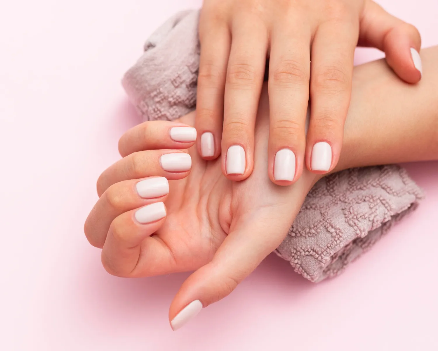 Manos con uñas pintadas de color nude, sobre una superficie rosa, con una toalla suave en el fondo. Ideal para temas de cuidado de uñas y belleza.